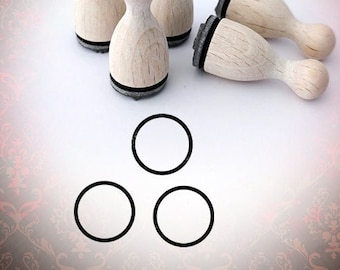 May include: Five wooden stamps shaped like bowling pins with black ink pads. The stamps are arranged in a semi-circle on a white surface. Three circles are drawn below the stamps.