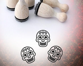 May include: Five wooden stamps with black ink pads, shaped like a rounded rectangle. The stamps feature a design of a sugar skull with floral details. The stamps are arranged on a white surface with a red and gold patterned background.