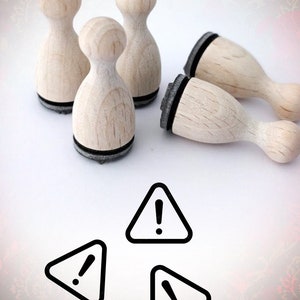 May include: Five wooden stamps with black ink pads. The stamps are shaped like a rounded rectangle with a rounded top. The stamps are arranged in a circle on a white surface. Three black warning signs with an exclamation point are in the center of the image.