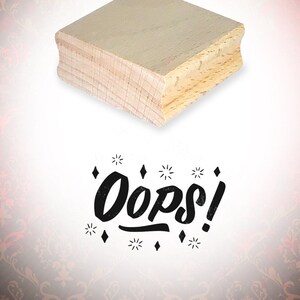 Könnte beinhalten: Ein Holz-Stempel mit dem Text "Oops!" in einer stilisierten Schriftart mit Diamanten drumherum.