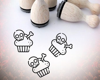 May include: Five wooden stamps with black ink pads, each featuring a skull and crossbones on a cupcake design. The stamps are arranged on a white surface with a red and gold patterned background.