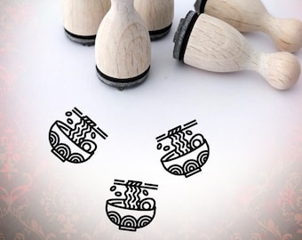 May include: Four wooden stamps with black ink pads, each featuring a line drawing of a bowl of ramen noodles with chopsticks. The stamps are arranged on a white surface with a red and gold patterned background.