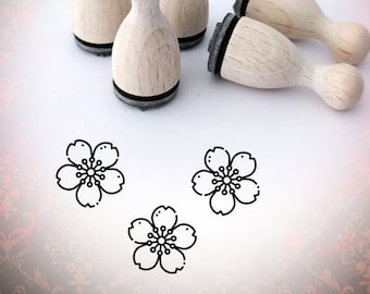 May include: Five wooden stamps with black ink pads, shaped like a rounded rectangle, with a flower design on the top. Three stamps are standing upright, and two are lying on their sides. Three black and white flower designs are visible on a white surface.