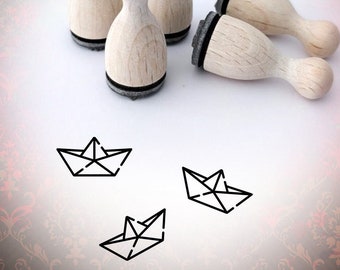 May include: Wooden stamps with a black ink pad base and a rounded handle. The image also shows three line drawings of paper boats. The background has a damask pattern with the text "Bloody Nose" in a cursive font.