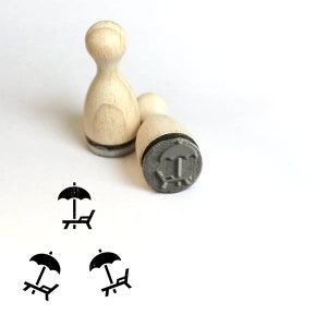 Puede incluir: Dos sellos de madera con almohadillas de tinta negra. Los sellos presentan un diseño de una silla de playa con una sombrilla.