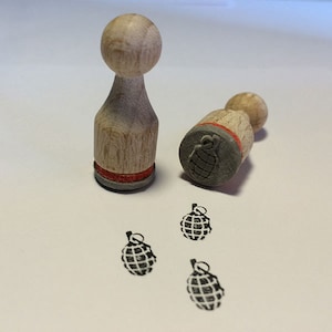 May include: Two wooden stamps with a red band around the top. The stamps are shaped like a small, round, wooden person. The stamps have a design of a hand grenade on the bottom. Three hand grenade stamps are also shown on a white surface.