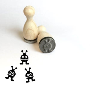 Könnte beinhalten: Drei Stempel aus Holz mit einem schwarzen und weißen Alien-Design. Die Stempel sind rund und haben einen Holzgriff. Das Alien-Design ist einfach und zeigt zwei Augen, einen Mund und zwei Antennen.