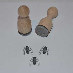 May include: Two wooden stamps with black rubber insets, each featuring a spider design. Three spider impressions are visible on the white surface below the stamps.