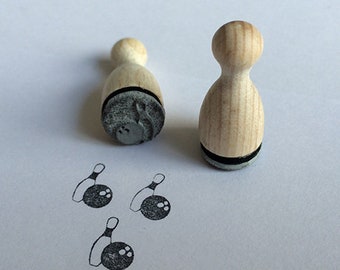 May include: Two wooden stamps shaped like bowling pins. The stamps are uninked and show the image of a bowling pin and ball. Three bowling pins and balls are also drawn on the white surface.