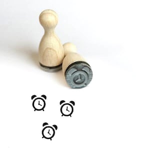 以下が含まれることがあります： 黒い目覚まし時計のデザインが施された木製のスタンプが3つ。スタンプは白い表面の上に置かれています。