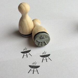 Könnte beinhalten: Vier Holzstempel mit Grillmotiv. Die Stempel sind bowlingpinförmig und haben ein graues Stempelkissen. Die Stempel befinden sich auf einer weißen Oberfläche.