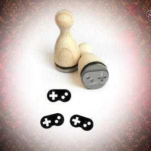 May include: Four wooden stamps with black ink pads. Three of the stamps are imprinted with a black and white gamepad design. The fourth stamp is a blank wooden stamp.