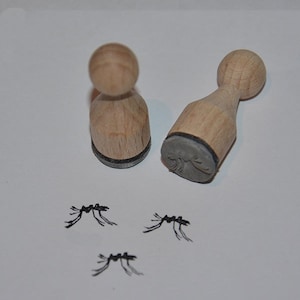 Pode incluir: Dois carimbos de madeira com desenhos de formigas e três imagens de formigas carimbadas. Os carimbos têm uma pega redonda de madeira e uma base de carimbo de borracha cinzenta. As imagens carimbadas são pretas e mostram o corpo e as pernas da formiga.