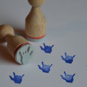 Könnte beinhalten: Zwei Holzstempel mit einem Handdesign. Die Stempel werden verwendet, um blaue Handabdrücke auf einer weißen Oberfläche zu erstellen. Die Handabdrücke haben die Form des deutschen Gebärdensprachzeichens für "Ich liebe dich".