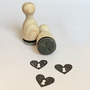 Könnte beinhalten: Drei Holzstempel mit schwarzen Stempelflächen. Die Stempel sind herzförmig mit einem Schlüsselloch-Design. Die Stempel befinden sich auf einer weißen Oberfläche und wurden verwendet, um drei herzförmige Abdrücke zu erstellen.