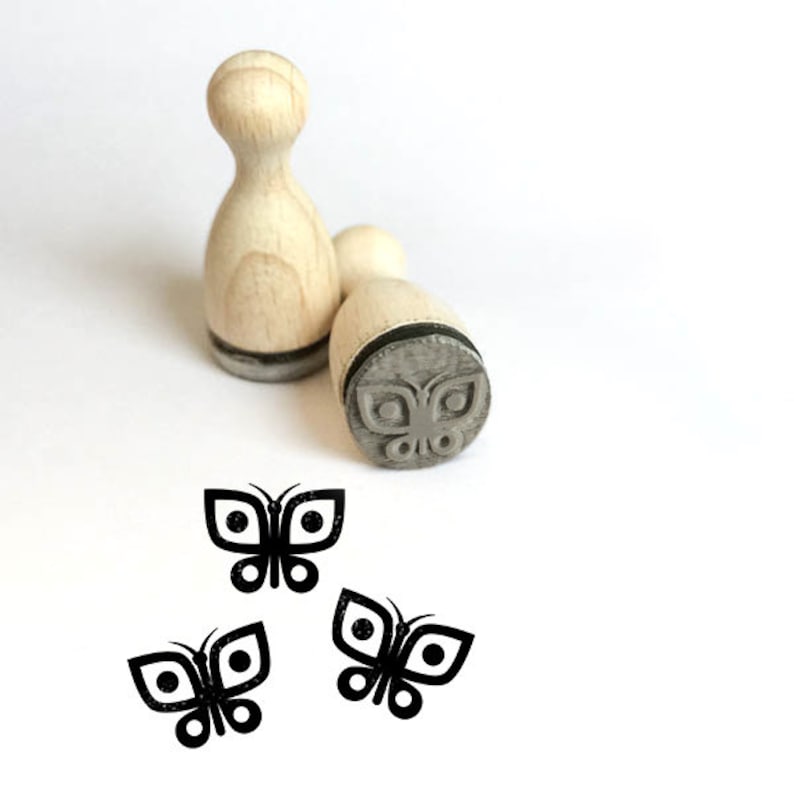 May include: Two wooden stamps featuring a butterfly design. The stamps are made from natural wood and have a black rubber base. The butterfly design is carved into the rubber base and is visible on the stamps. The stamps are ideal for adding a touch of whimsy to your crafts.