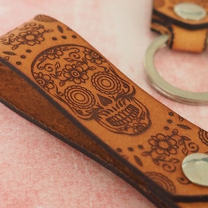 May include: A brown leather keychain with a silver key ring and a laser-etched sugar skull design. The skull is surrounded by floral patterns.
