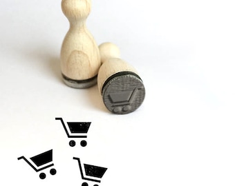 May include: Two wooden stamps with a shopping cart design. One stamp is upright, and the other is lying on its side, showing the shopping cart image. Three black shopping cart icons are printed below the stamps.