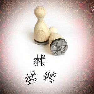 Könnte beinhalten: Zwei Holzstempel mit einer grauen Gummioberfläche. Die Stempel sind abgerundet und haben einen schwarzen Streifen am Boden. Die Gummioberfläche der Stempel hat ein Tic-Tac-Toe-Spieldesign. Die Stempel befinden sich auf einer weißen Oberfläche mit einem roten und goldenen Muster im Hintergrund.