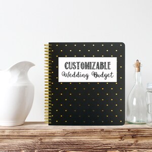Op de afbeelding: Een zwart spiraalgebonden notitieboek met gouden stippen en een gouden spiraal. Het notitieboek heeft de tekst "CUSTOMIZABLE Wedding Budget" op een witte rechthoek. Een witte kan, een glazen fles en een pot staan op een houten oppervlak.