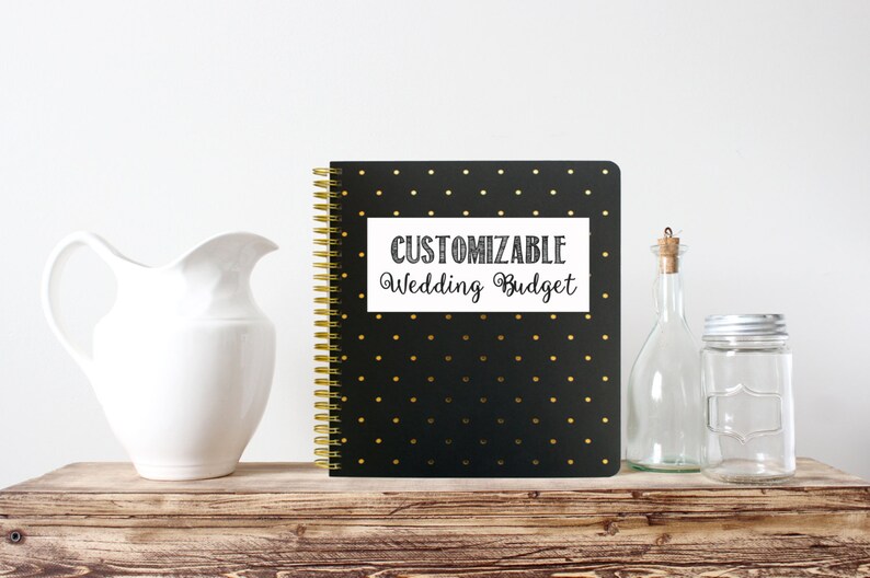 Op de afbeelding: Een zwart spiraalgebonden notitieboek met gouden stippen en een gouden spiraal. Het notitieboek heeft de tekst "CUSTOMIZABLE Wedding Budget" op een witte rechthoek. Een witte kan, een glazen fles en een pot staan op een houten oppervlak.