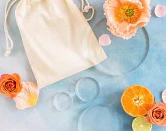 May include: A white cotton drawstring bag with a light blue background. The bag is surrounded by orange and pink flowers, a sliced orange, a lime, and clear glass circles.