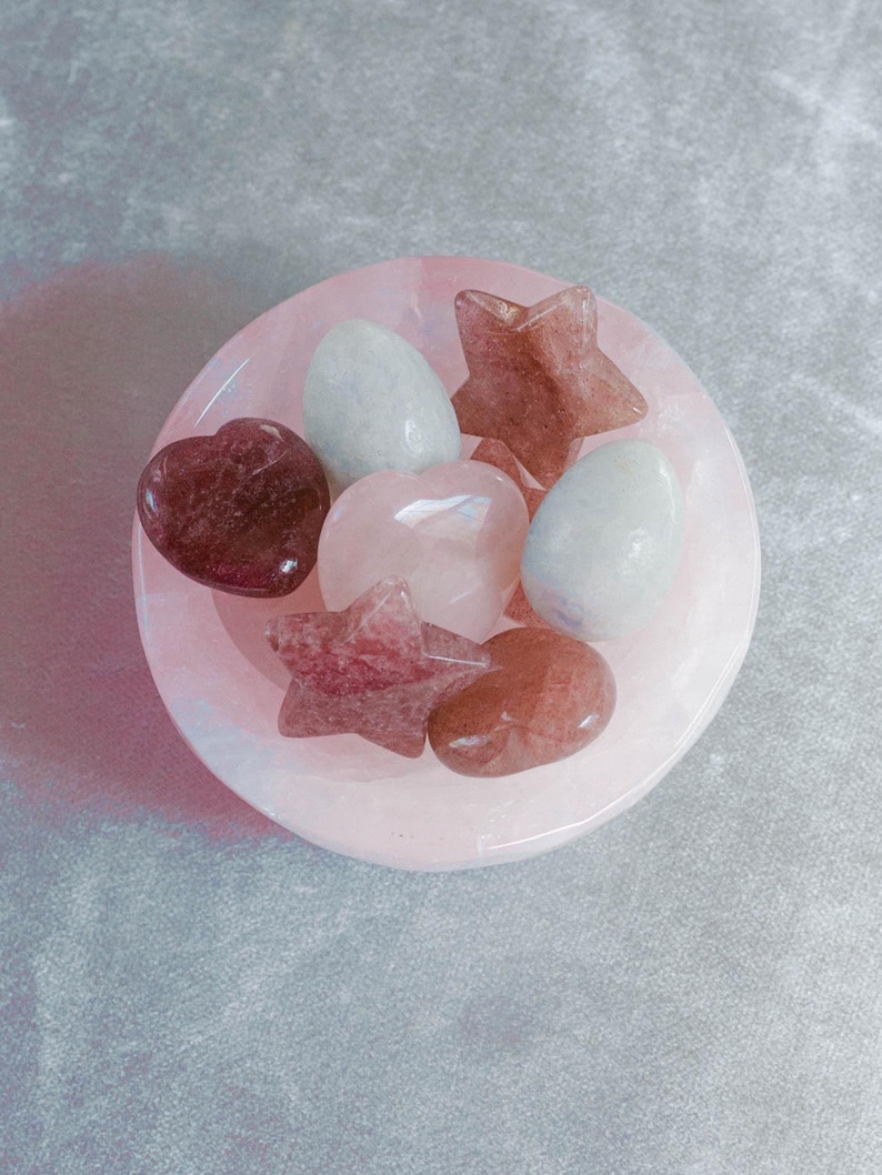 A polished bowl of mixed healing crystals in soft pink, red, and pale blue tones, including rose quartz hearts, strawberry quartz stars, and blue calcite stones, displayed on a textured gray background.