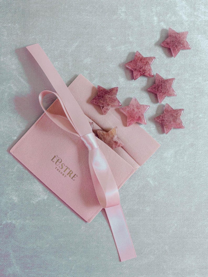 May include: A pink velvet pouch with the text "L'ESTRE THEORY" and a pink satin ribbon. Scattered around the pouch are six star-shaped, pink gemstones. The gemstones have a speckled appearance and are arranged on a light blue-gray surface.