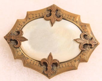 May include: An antique oval-shaped brooch with a gold-toned frame and a mother-of-pearl center. The frame features a decorative pattern and fleur-de-lis accents at each corner and the center.