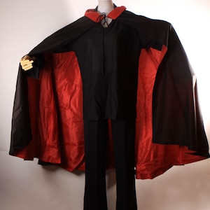 May include: A black cape with a red lining and a matching black outfit. The cape has a red collar and is displayed on a mannequin. The outfit includes a black shirt and black pants.