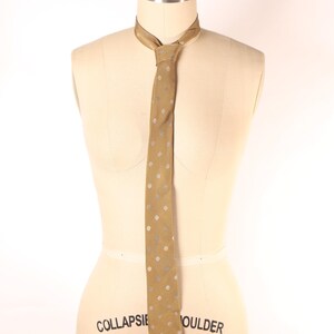 Peut inclure: Une cravate dorée avec un motif répété de petits points clairs. La cravate est présentée sur un mannequin. La cravate est nouée au cou. Le mannequin porte l'inscription "COLLAPSIBLE SHOULDER".