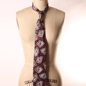 May include: A maroon tie with a repeating geometric pattern of white and blue squares and diamond shapes. The tie is displayed on a mannequin. The tie is knotted at the neck.