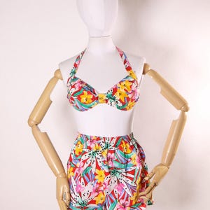 Puede incluir: Un top de bikini y pantalones cortos a juego con estampado floral rojo, blanco, azul, verde y amarillo. El top de bikini tiene un estilo halter y los pantalones cortos tienen cintura alta.