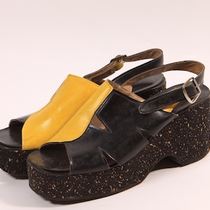 May include: A pair of black and yellow platform sandals with cork wedges. The sandals have a strap across the foot and an ankle strap with a buckle.