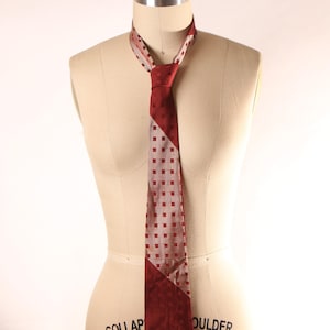 May include: A burgundy and gray patterned tie displayed on a mannequin. The tie features a geometric design with squares and diagonal color blocking. The tie is a classic accessory for formal wear.