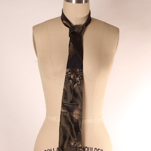 Puede incluir: Corbata estampada en verde oscuro y marrón expuesta sobre un maniquí. La corbata presenta un diseño geométrico y floral con toques de color beige y negro. La corbata está anudada al cuello.