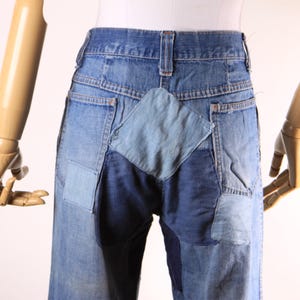 May include: Blue denim jeans with patchwork detailing. The jeans feature a light blue diamond-shaped patch on the back, with other patches in varying shades of blue. The jeans are displayed on a mannequin.