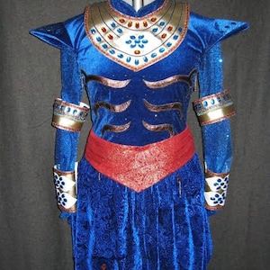Puede incluir: Un disfraz azul con detalles dorados y rojos. El disfraz presenta un cuello decorativo, hombreras y detalles intrincados en las mangas y el corpi&ntilde;o. Un cintur&oacute;n rojo estampado y pantalones azules completan el look.