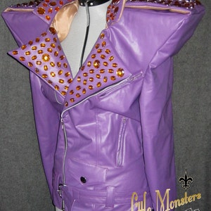 以下が含まれることがあります： 紫色のフェイクレザーのジャケット。大きな肩パッドとノッチドラペルが付いています。オレンジ色のラインストーンが多数あしらわれています。シルバーのジッパーとウエストベルトが付いています。「Little Monsters Couture」の文字が見えます。