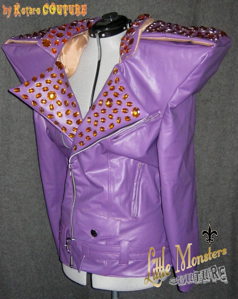 以下が含まれることがあります： 紫色のフェイクレザーのジャケット。大きな肩パッドとノッチドラペルが付いています。オレンジ色のラインストーンが多数あしらわれています。シルバーのジッパーとウエストベルトが付いています。「Little Monsters Couture」の文字が見えます。