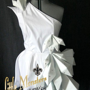 以下が含まれることがあります： 幾何学的なアクセントが施された白いワンショルダーのドレス。 ドレスは、フィットしたボディスとフレアスカートが特徴です。 デザインには、フルール・ド・リスのエンブレムと「Little Monsters FUTURE by Kotaro COUTURE」の文字が含まれています。