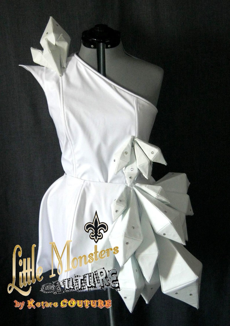 以下が含まれることがあります： 幾何学的なアクセントが施された白いワンショルダーのドレス。 ドレスは、フィットしたボディスとフレアスカートが特徴です。 デザインには、フルール・ド・リスのエンブレムと「Little Monsters FUTURE by Kotaro COUTURE」の文字が含まれています。