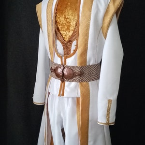 Puede incluir: Un disfraz blanco con detalles dorados, con cuello alto y mangas largas. El atuendo incluye un cintur&oacute;n decorativo y pantalones a juego. El disfraz es probablemente para una actuaci&oacute;n teatral o cosplay.