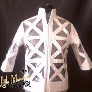 May include: White jacket with geometric cutouts. The jacket has a high collar and a full-length zipper. The sleeves are three-quarter length. The jacket is displayed on a mannequin. The text "Little Monsters Couture" is in the lower left corner.