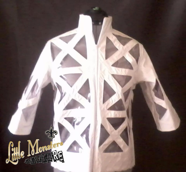 May include: White jacket with geometric cutouts. The jacket has a high collar and a full-length zipper. The sleeves are three-quarter length. The jacket is displayed on a mannequin. The text "Little Monsters Couture" is in the lower left corner.