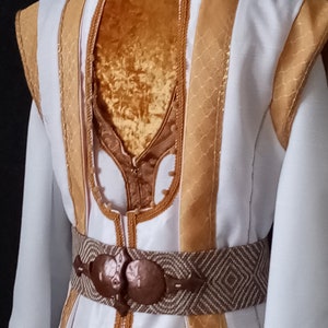 Puede incluir: Un disfraz blanco y dorado con cuello alto y adornos decorativos. El disfraz presenta detalles dorados en los hombros, las mangas y el corpi&ntilde;o. Un cintur&oacute;n marr&oacute;n estampado con hebilla de bronce ci&ntilde;e la cintura. La parte trasera del disfraz es visible.