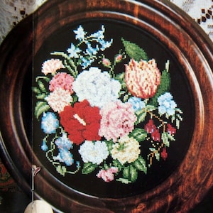 May include: A framed cross-stitch embroidery depicting a colorful floral arrangement with pink, red, white, and blue flowers. The flowers are arranged in a circular pattern on a black background. The frame is made of dark wood with a rounded edge.