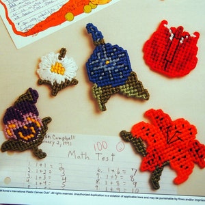 May include: A collection of colorful plastic canvas flower fridge magnets. The magnets are in the shapes of a sunflower, a daisy, a tulip, a poppy, and a bluebell. The magnets are all handmade and feature a variety of colors and patterns.