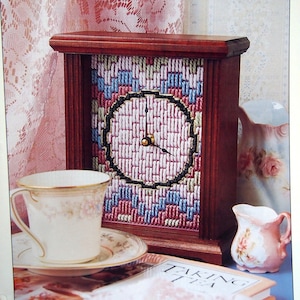 Puede incluir: Un reloj de madera con un diseño bargello en tonos de rosa, azul y verde. La esfera del reloj es un círculo con agujas negras y un centro dorado. El reloj está sentado en una mesa con una taza y un platillo, una jarra y un libro.