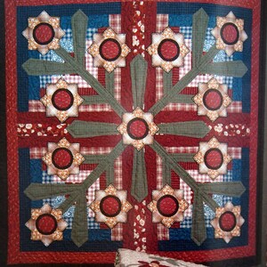 Op de afbeelding: Een rood, wit en blauw patchwork-deken met een madeliefje-motief. Het deken is gevouwen en geplaatst in een rieten mand met een label met de tekst "Daisy Bouquet".